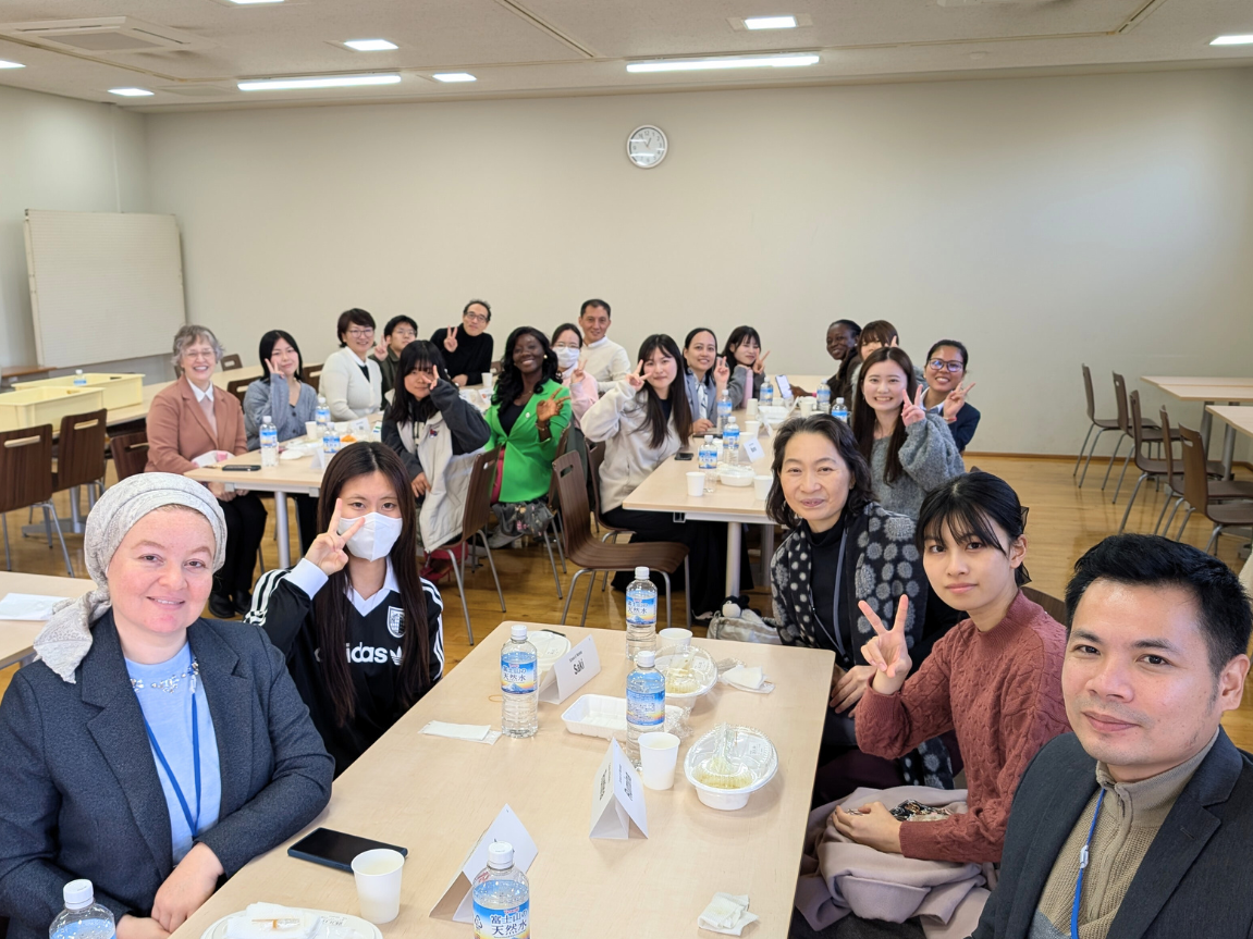 8か国の研修員と本学学生が笑顔でピースサインをしながら集合写真を撮っている様子。テーブルには名札やペットボトル、食事の容器が並び、和やかな交流の雰囲気が伝わる。